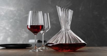 Die mit Rotwein gefüllte SPIEGELAU Lifestyle Karaffe auf einem Tisch. Links von der Karaffe zwei Lifestyle Rotweingläser, eines davon mit Rotwein gefüllt. Am linken Rand des Tisches eine leere schwarze Platte.<br/>