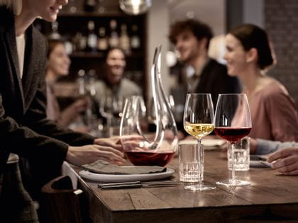 The filled white wine glass and the filled Bordeaux glass SPIEGELAU Willsberger Anniversary in front of the SPIEGELAU Perfect Serve Collection SOF glass and the filled SPIEGELAU Novo decanter. All of them on a dinner table surrounded by people.<br/>